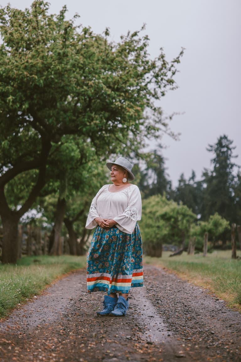 A woman in a ribbon skirt standing on a wet orchard lane