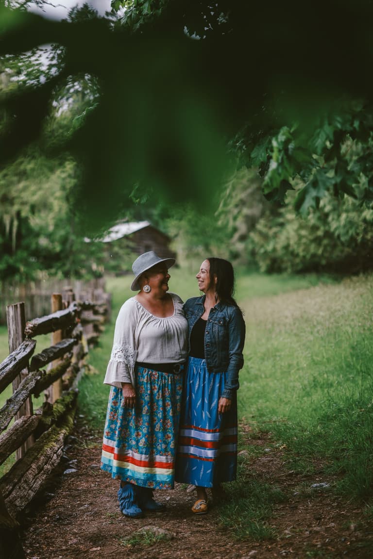 Two women in ribbon skirts beside a split rail fence