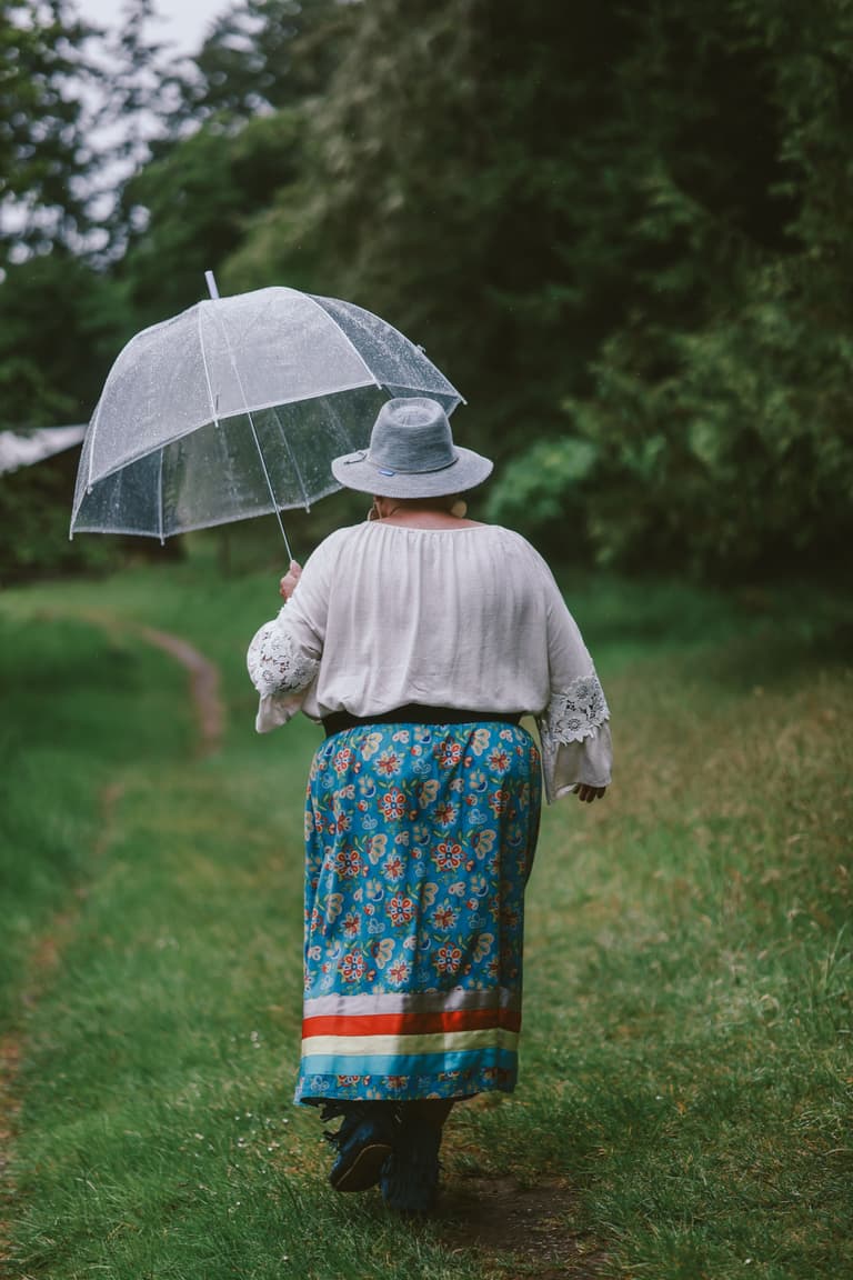 A blue ribbon skirt with red and white satin bands, walking an orchard path under a clear umbrella