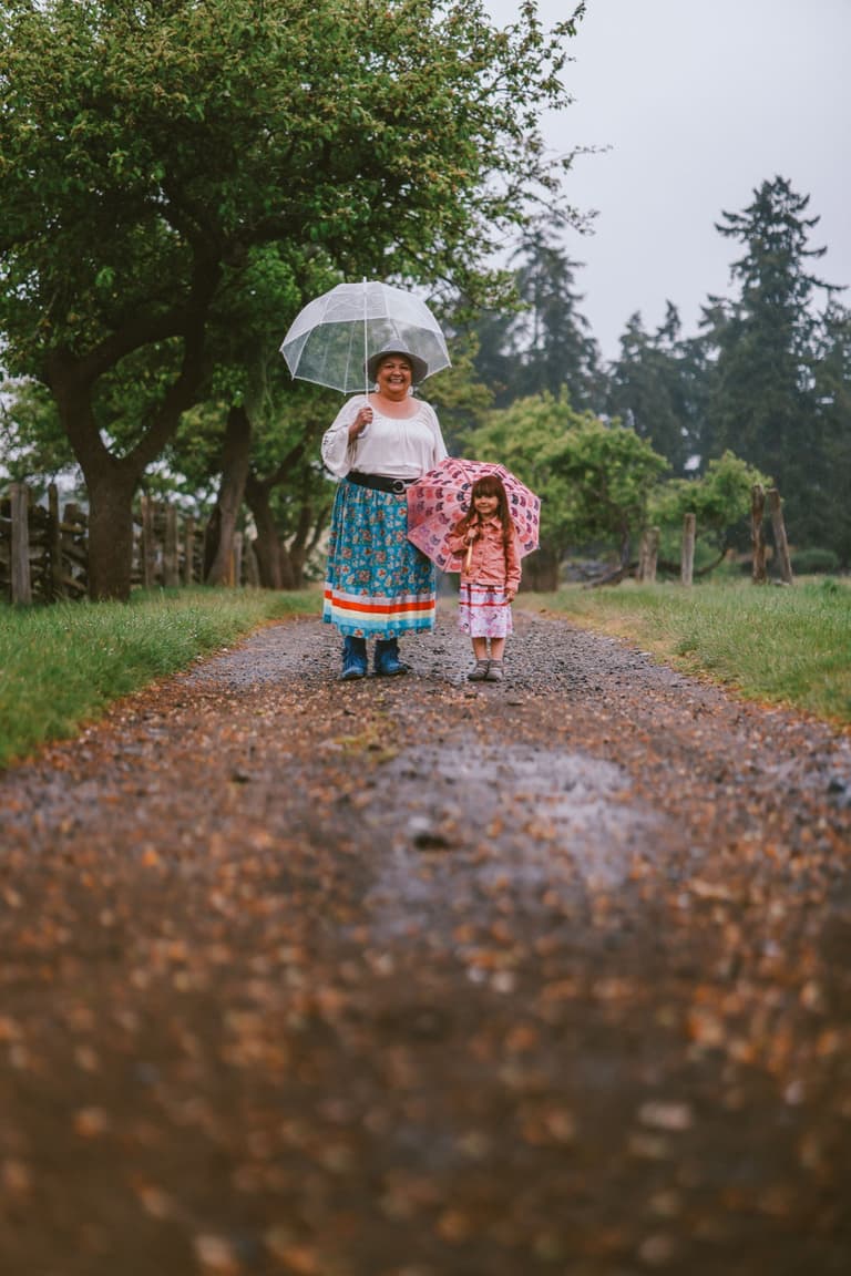 A woman and child walking a wet orchard path under a clear umbrella