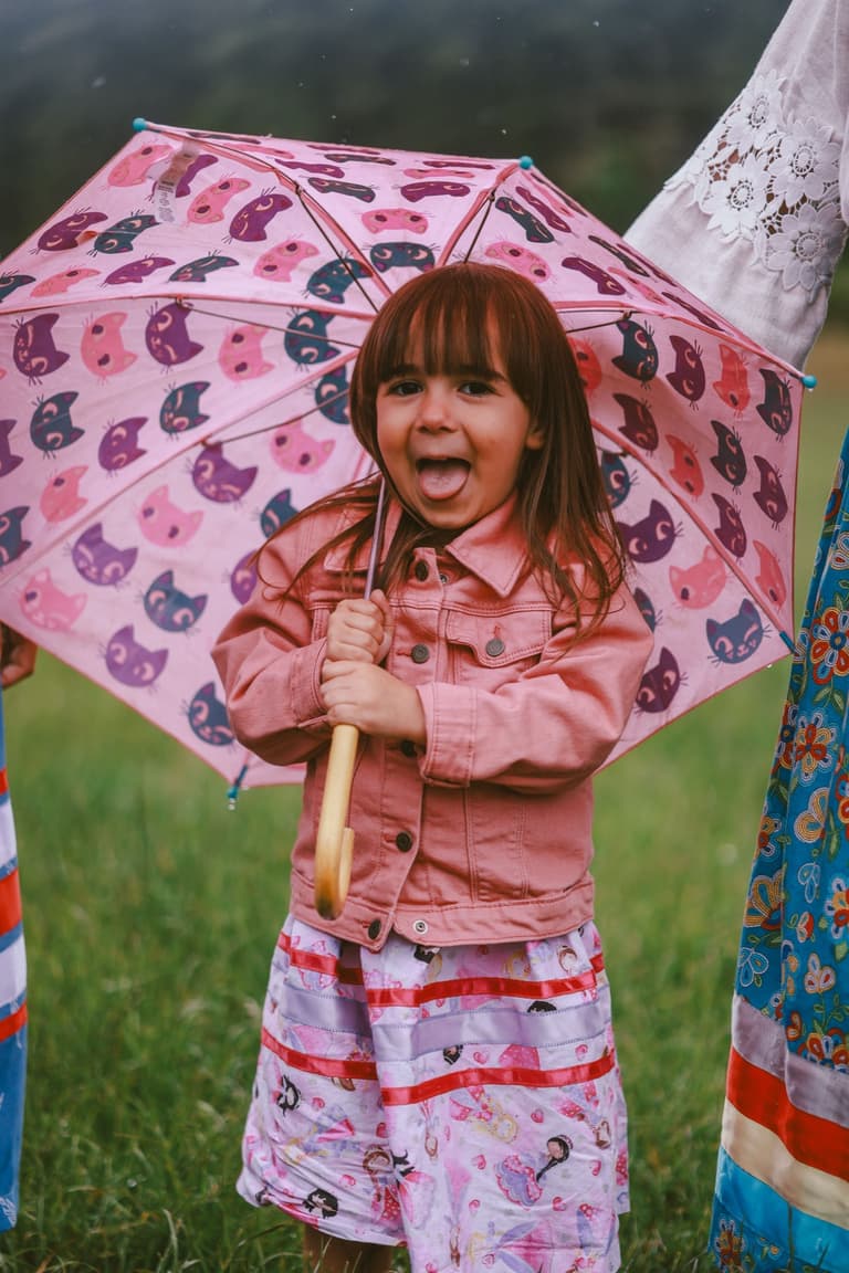 A young girl laughing under a patterned umbrella, wearing a ribbon skirt
